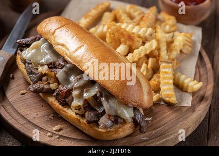 Sandwich di cheesesteak di Philly preparato con bistecche, formaggio e cipolle su un rotolo di hoagie con patatine fritte su un asse di legno Foto Stock