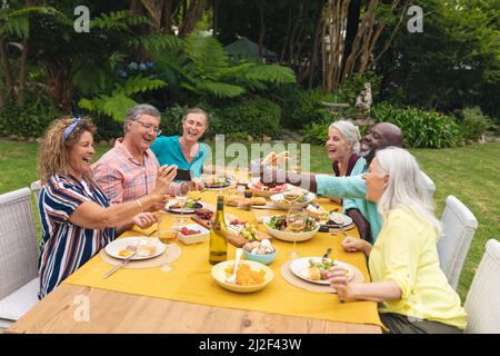 Amici senior multirazziali che guardano lo smartphone mentre mangiano durante la festa del cortile Foto Stock