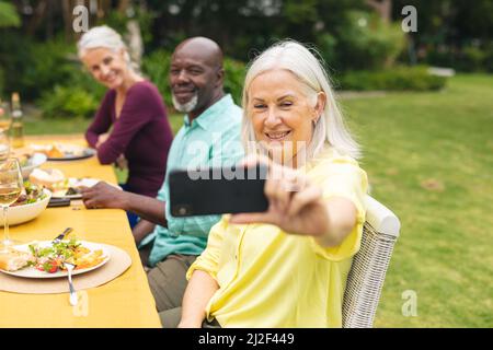 Donna anziana che prende selfie con amici multirazziali che hanno cibo al partito del cortile Foto Stock