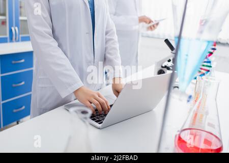 visualizzazione ritagliata della digitazione da parte dello scienziato su un laptop in laboratorio vicino a un collega sfocato,immagine stock Foto Stock