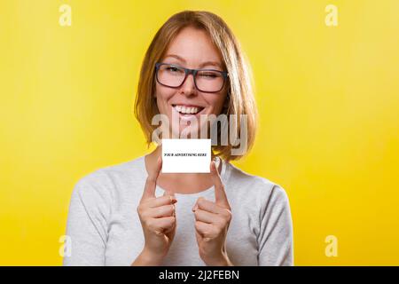 Affari e pubblicità. La giovane donna bionda in occhiali, tiene indice dita entrambe le mani carta bancaria o biglietto da visita. Sfondo giallo. Copia sp Foto Stock