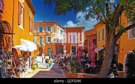 Roussillon en Provence, Francia - Ottobre 2. 2019: Vista oltre l'albero sulla piazza, giallo brillante, ocra e rosso case mediterranee, persone seduti outsid Foto Stock