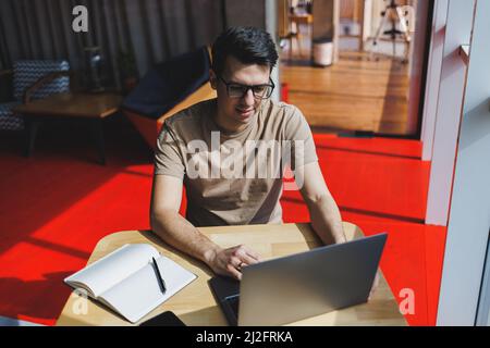 Giovane maschio millenario seduto al tavolo davanti aprire computer portatile mentre studiano online. Moderno nomade digitale lavora su freelance su netbook in coffee Foto Stock