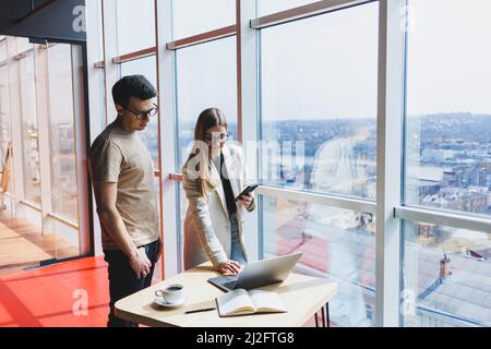 Due colleghi, un uomo e una donna manager, stanno accanto a un tavolo con un laptop in un ufficio moderno e discutono di un nuovo progetto. Due partner commerciali in gla Foto Stock