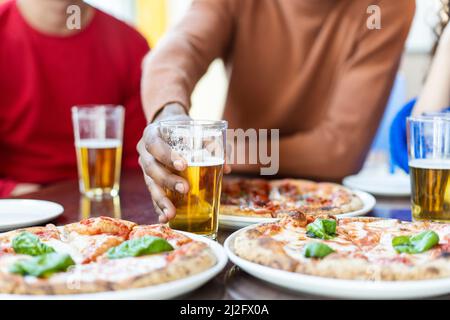 Primo piano del ragazzo africano che prende a mano un bicchiere di birra al ristorante pizza - amici multietnici che si divertono a mangiare pizza e bere birra al pub Foto Stock