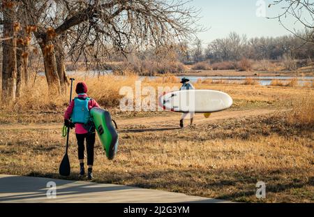 Evans, CO, Stati Uniti d'America - 26 marzo 2022: Le pagaiole femminili stanno trasportando il loro stand gonfiabile su paddleboards per lanciare per il primo viaggio primaverile di pagaioli sulla S Foto Stock