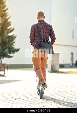 Giovane uomo in pantaloni casual e maglietta che guida il suo scooter elettrico, guarda la forma dietro, edifici sullo sfondo Foto Stock