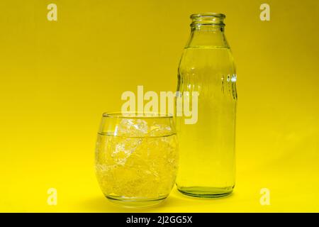 Bicchiere di vetro e bottiglia riempiti di ghiaccio e acqua depurata. Sfondo giallo. Foto Stock