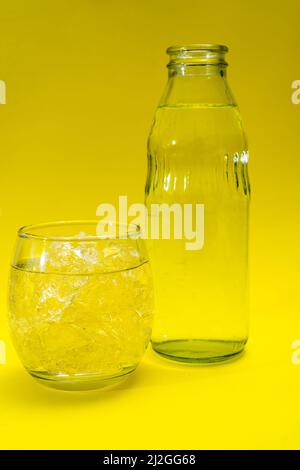 Bicchiere di vetro e bottiglia riempiti di ghiaccio e acqua depurata. Sfondo giallo. Foto Stock