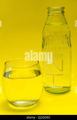 Bicchiere di vetro e bottiglia riempiti con acqua purificata. Sfondo di Yelloew. Foto Stock