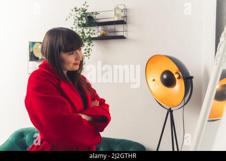 Donna cliente europea sicura che guarda se stessa nello specchio mentre prova il cappotto rosso d'inverno. Foto di alta qualità Foto Stock