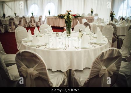 Una sala per matrimoni vuota con tavoli rotondi e puliti ricoperti di tovaglie bianche e decorati con fiori Foto Stock