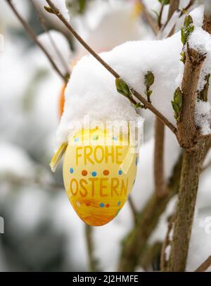 02 aprile 2022, Hessen, Francoforte sul meno: "Buona Pasqua" è scritto su un uovo di Pasqua appeso ad un arbusto in un cortile di fronte, coperto di neve, dopo che ha nevicato il giorno prima e la notte prima. Foto: Frank Rumpenhorst/dpa Foto Stock