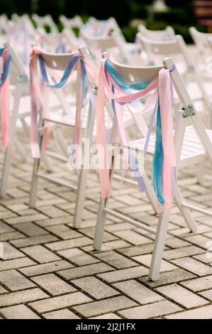 Sedie di legno bianche decorate con nastri rosa e blu. Cerimonia nuziale all'aperto Foto Stock