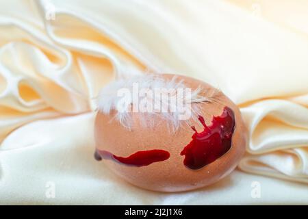 un uovo di pollo con una piuma e un sangue come vernice, su pieghe beige satinato Foto Stock