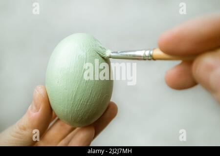 La mano tiene il guinzaglio e dipinge l'uovo. Un processo di pittura di un uovo di Pasqua con vernice verde chiaro. Pennello in mano della donna su sfondo grigio. Tradizione Foto Stock