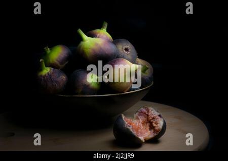 Chiaroscuro, fotografia in stile barocco rinascimentale ancora vivo, fichi in una ciotola su un tagliere e un fico tagliato fuori dalla ciotola, sfondo nero, Foto Stock