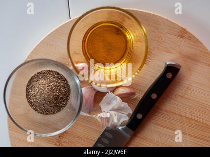 Un bicchiere con olio d'oliva e un bicchiere con pepe nero con aglio e un coltello su un tagliere di legno, come parte degli ingredienti per la cottura. Vista dall'alto Foto Stock