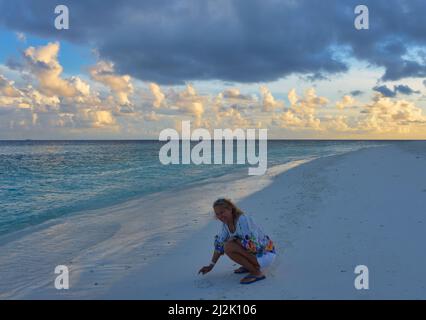 Donna sorridente accoccolata sulla spiaggia che si disegna nella sabbia all'alba, Maldive Foto Stock