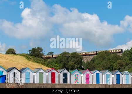 Case colorate sulla spiaggia. Fila di capanne multicolore sulla spiaggia con treno a vapore vintage soffiato su viadotto di pietra contro il cielo blu. Foto Stock