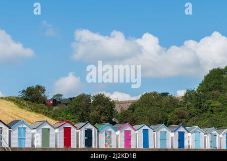 Case colorate sulla spiaggia. Fila di capanne multicolore sulla spiaggia con treno a vapore vintage soffiato su viadotto di pietra contro il cielo blu. Foto Stock