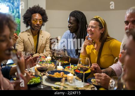 Persone diverse di età ed etnia che mangiano una cena vegana. Gruppo multietnico di amici divertirsi mentre si condivide un pasto in un caldo e accogliente hous Foto Stock