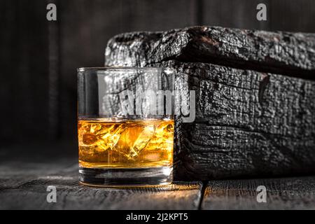 Ghiaccio con whisky dorato in vetro su legno nero bruciato. Whisky su rocce su tavola bruciata. Foto Stock
