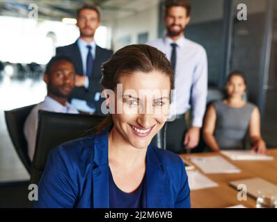 Fidatevi quando tutti gli altri dubitano di voi. Ritratto di una donna d'affari sorridente seduta in un ufficio con i colleghi sullo sfondo. Foto Stock