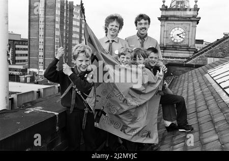 Volando la bandiera... questo è ciò che sta facendo il pack Netherton Moor Cub per festeggiare il 75th compleanno del movimento - e visto con la bandiera del gruppo sono (davanti, da sinistra) nove anni gemelli Edward e James Broadbent e fratelli Edward, 10 e Gareth Johns, otto. Nella foto sono raffigurati anche i genitori e i capi Cub Susan e Kevin Johns. Ai cubani di tutto il paese è stato chiesto di progettare la propria bandiera e poi trovare luoghi insoliti per volare loro. La bandiera Netherton viene volata dall'edificio Co-op di New Street, una delle più alte del centro della città di Huddersfield. 24th giugno 1991. Foto Stock