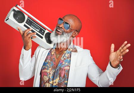 Il tipo di musica che ascolti dice molto su di te. Studio girato di un uomo anziano indossando abiti vintage mentre si posa con un boombox contro un rosso Foto Stock