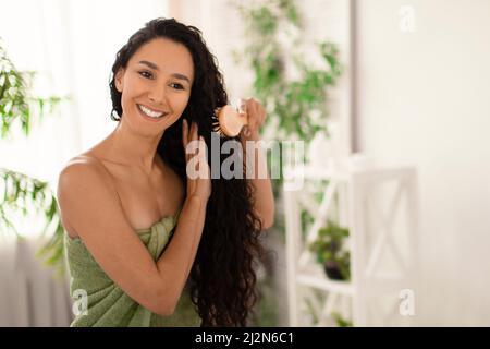 Bella ragazza felice in asciugamano spazzolando i suoi capelli lunghi ondulati con spazzola di fronte allo specchio in bagno, spazio libero Foto Stock