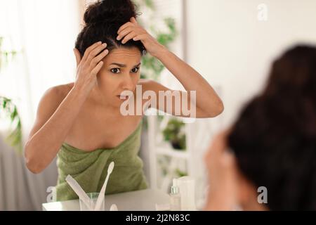 Preoccupata donna millennial che ha il problema di dandruff o capelli grigi, guardando le sue radici nello specchio al bagno Foto Stock