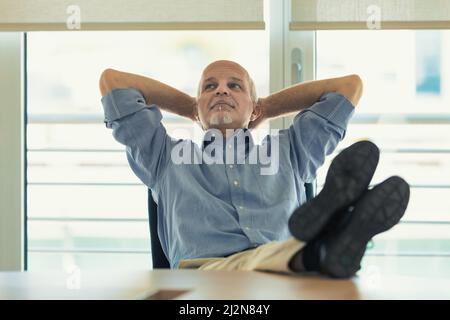 L'uomo in ufficio si rilassa con le braccia incrociate dietro la testa e i piedi sul tavolo, sognando la vacanza, il fine settimana o la pensione Foto Stock