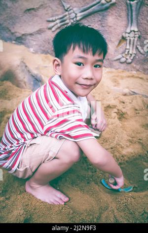 Bambino felice che gioca con la sabbia e sorridente. Adorabile ragazzo asiatico ha divertimento scavando nella sabbia in una giornata estiva e guardando la macchina fotografica. Tono vintage. All'aperto Foto Stock