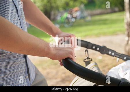 Un giovane papà che cammina nel parco con un bambino in un passeggino. Primo piano delle mani degli uomini con un passeggino. Concetto di Father's Day. Foto di alta qualità Foto Stock