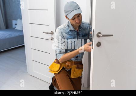 Montaggio di una serratura sulla porta di ingresso in legno anteriore. Ritratto di giovane fabbro operaio in uniforme blu montaggio pomello porta. Professionale Foto Stock