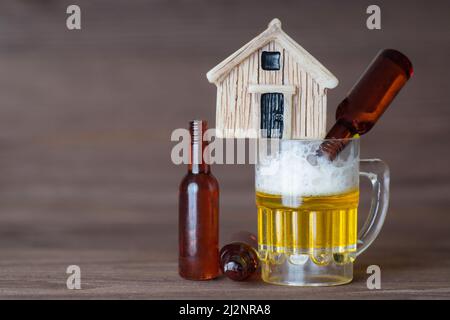 Casa giocattolo in miniatura in piedi su una piccola tazza di birra con bottiglie vuote che si trovano intorno. Alcolismo e problemi familiari. Foto Stock
