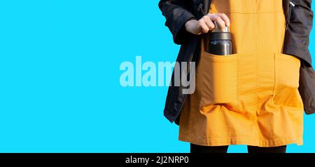 Modello femminile con un impermeabile nero su un abito arancione e un pallone thermos in una tasca isolata su sfondo blu. Foto Stock