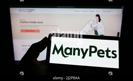 Persona che detiene il telefono cellulare con il logo della compagnia di assicurazione acquistato da Many Ltd. (ManyPets) sullo schermo di fronte alla pagina web. Mettere a fuoco sul display del telefono. Foto Stock