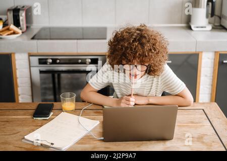 Giovane studentessa in auricolari utilizzando il cellulare mentre studiava con il computer portatile in cucina a casa, pensando Foto Stock