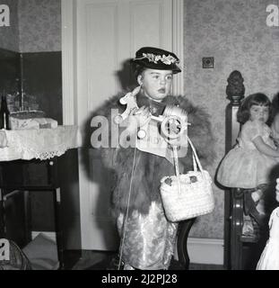 1961, storica, in un abito lucido, top in pelliccia, cappello fantasia, gioielli e borsa a spalla, una giovane ragazza in piedi su una sedia, vestita per il suo compleanno come 'Lady Docker', Stockport, Manchester, Inghilterra, Regno Unito. Nel dopoguerra britannico, Lady Docker - nato Norah Royce Turner in un appartamento sopra un macellaio a Derby - era una 'personalità', una socialite. Attraverso il matrimonio, in particolare il suo terzo matrimonio con Sir Bernard Docker, presidente della Daimler e direttore della Midland Bank, ha condotto per il tempo una vita colorata e stravagante, ma non ha mai dimenticato le sue umili radici della classe operaia. Foto Stock