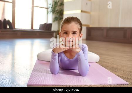 Bambina che indossa un abito viola . Ragazza sorridente carina che si trova sul pavimento su un tappeto sportivo Foto Stock