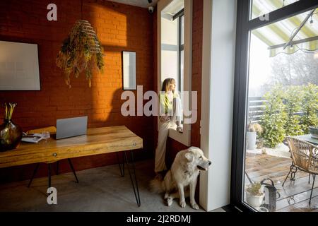 Donna seduta sul davanzale della finestra e guardando fuori la finestra con il suo cane bianco adorabile a casa Foto Stock