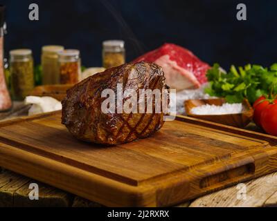 Cotto appetitoso prosciutto di carne su un tagliere di legno. Sullo sfondo si trova un pezzo di carne cruda, spezie, erbe, sale su sfondo blu scuro. Ricette Foto Stock