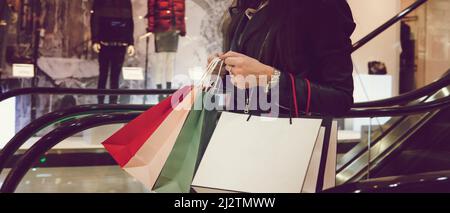 Scatto orizzontale di donna di raccolto in giacca nera di cuoio che tiene molti sacchetti di shopping e che si muove giù sulla scala mobile nel centro commerciale. Concetto di spesa in sa Foto Stock