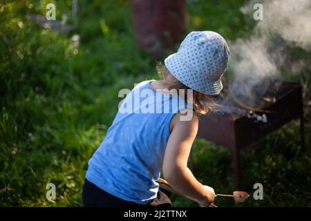 Bambini al picnic. La ragazza cuoce il pane sul bastone. Attività ricreative estive all'aperto. Bambino vicino al fuoco. Bambino a panama. La ragazza tiene il bastone di pane sopra il fuoco. Foto Stock