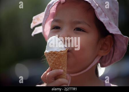 Il bambino sta mangiando il gelato. Ragazza che lecca il gelato per strada. Un cono di waffle nelle mani di un bambino asiatico. Ritratto di una ragazza carina con dolce Foto Stock