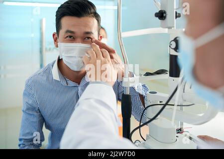 Uomo asiatico in maschera medica lamentarsi di dolore nell'occhio sinistro quando si parla con l'oftalmologo Foto Stock