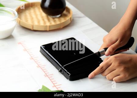 Donna Homemaker taglio Cincau Hitam o Black Grass Jelly Pudding in pezzi, cibo sano. Preparazione di Takjil per Buka Puasa Ramadhan Foto Stock