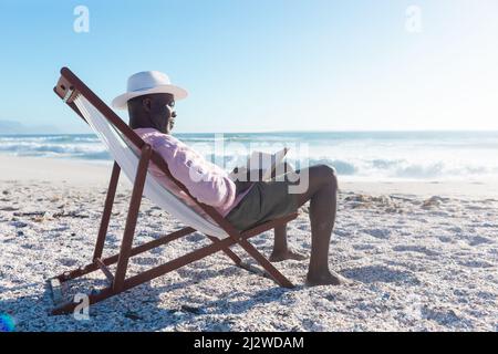 Vista laterale di un uomo anziano africano che legge libro seduto su sedia pieghevole in spiaggia Foto Stock